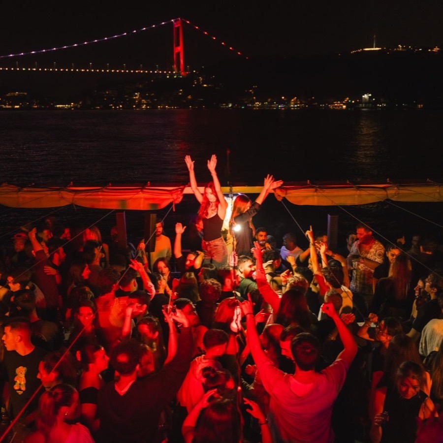 Crowd dancing on deck at night with the Bosphorus Bridge lit up red in the background